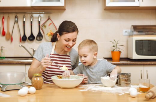 Little kid boy helps mother to cook Christmas ginger biscuit in light kitchen with tablet on the table. Happy family mom 30-35 years and child 2-3 in weekend morning at home. Relationship concept.
